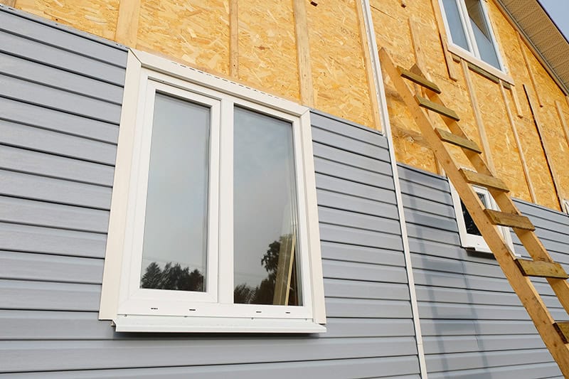 A house under construction with partially installed gray vinyl siding. The top half of the exterior wall features exposed insulation and wooden panels. A white-framed window is already in place, showcasing expert window installation Buchanan County, and a wooden ladder leans against the wall.