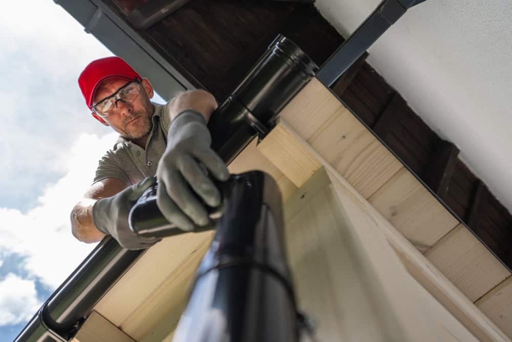 A man wearing a red hard hat, glasses, gloves, and a gray shirt is installing black rain gutters on a roof. He is positioned on the roof, working attentively with the gutter system against a backdrop of blue skies and white clouds. This scene exemplifies Gutter Services Buchanan County at its finest.