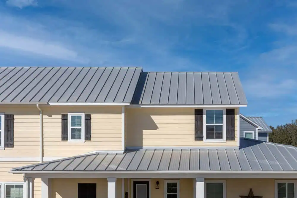 A yellow two-story house with a gray metal roof and white trim. The house features black window shutters, multiple windows, and a small front porch. The sky above is clear and blue, indicating a sunny day.