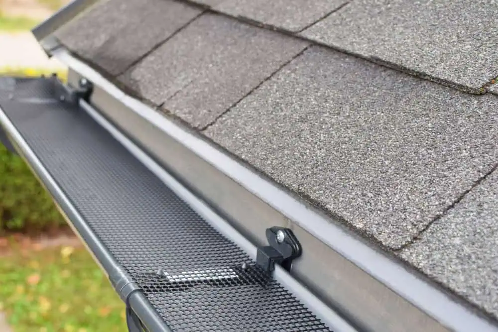 Close-up of a house roof with asphalt shingles and a black metal gutter guard installed along the edge. The gutter guard features a fine mesh cover designed to keep debris out of the gutter. Parts of a sloping roof and some greenery are visible in the background.