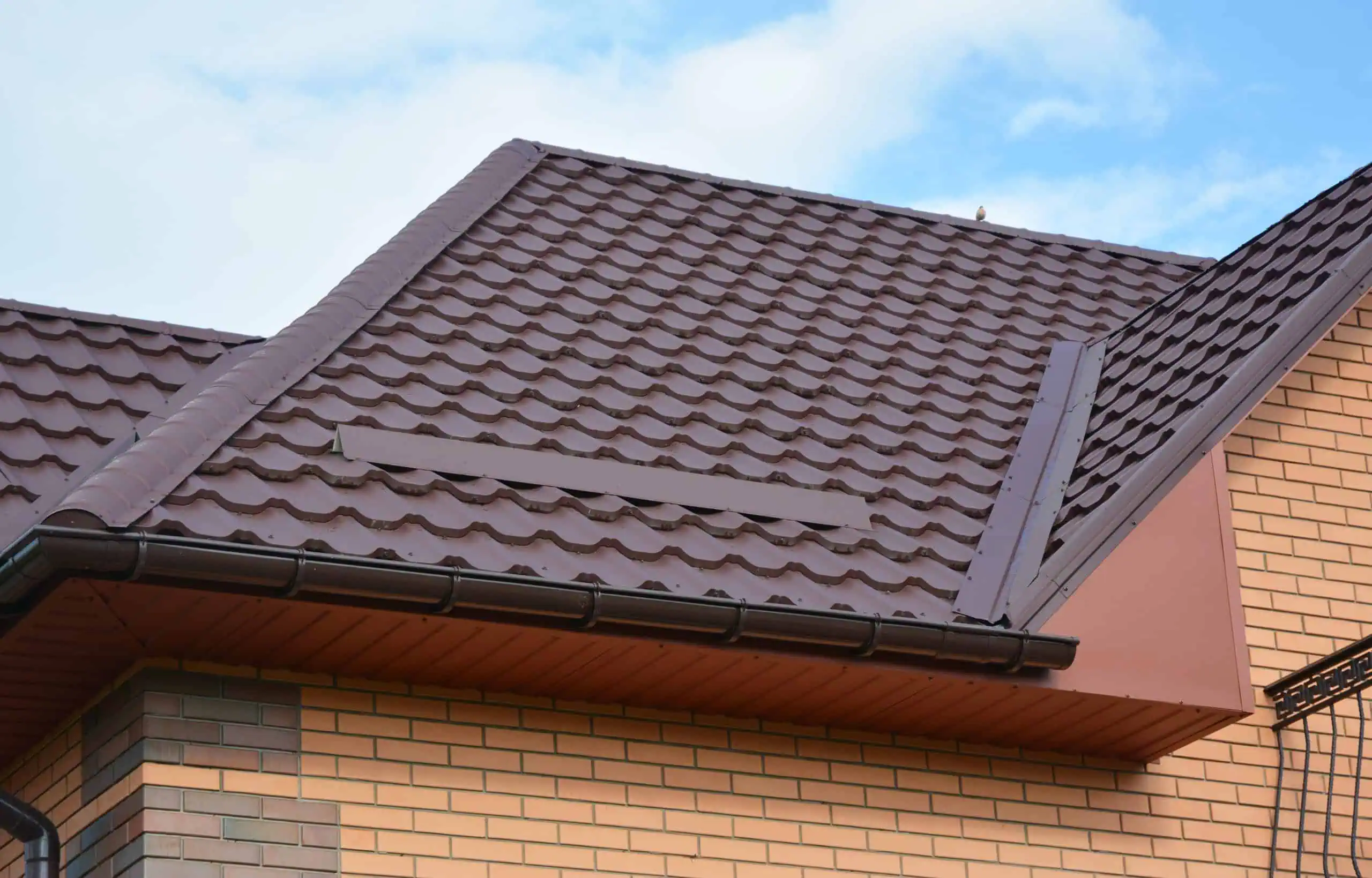 A close-up of a house roof with brown metal tiles, a dark gutter, and beige brick walls beneath, set against a partly cloudy blue sky.