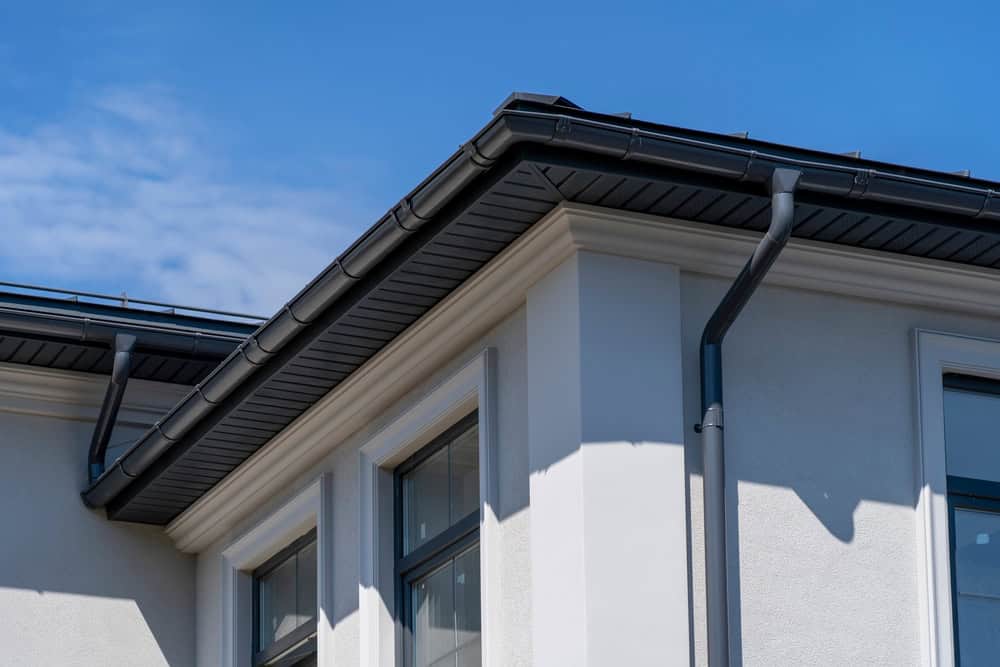 Close-up view of a modern house corner with white walls and large windows, showcasing black roof gutters and downspouts against a clear blue sky. The structure is well-defined with sharp lines and shadows, highlighting the building's contemporary design.