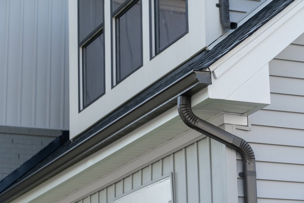 Close-up of a house exterior showing a multi-story section with large windows, siding, and a corner where a dark-colored downspout connects to a gutter. The upper part has horizontal siding, and the lower part has vertical siding. The structure appears modern.