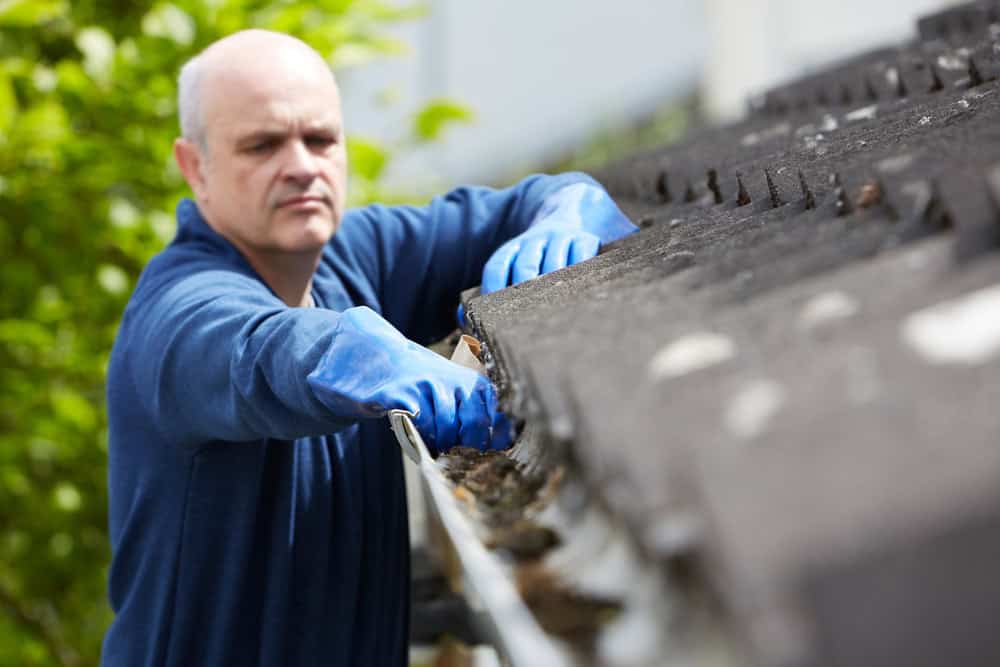 A man wearing blue gloves and a blue shirt is cleaning debris from a gutter attached to the roof of a house. The background is blurred, showing green foliage.