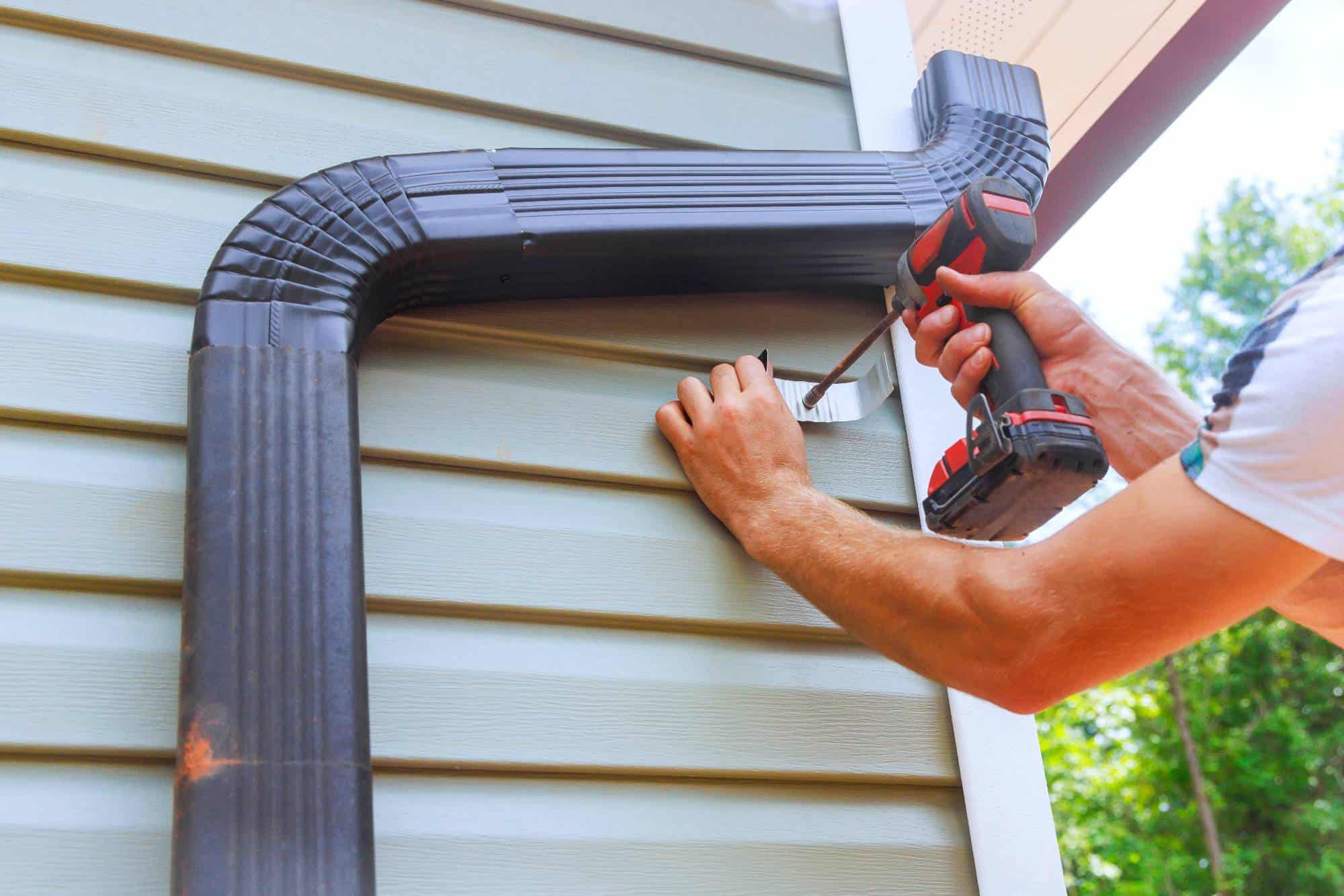A person uses a cordless power drill to secure a black downspout to the side of a house with light green siding. The downspout curves at a right angle near the roof. Trees are visible in the background.